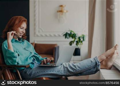 Relaxed female student with ginger hair dressed in shirt and jeans, keeps laptop on knees, browses necessary information, prepares for passing exams. Teacher conducts training lesson with pupils