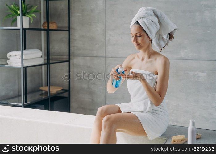 Relaxed latina woman applying skincare cosmetics sitting on bath in modern bathroom. Female wrapped in towel squeezing cosmetic product from bottle for body care treatment after shower.. Woman apply natural skincare cosmetics sitting in modern bathroom. Body care treatment after shower