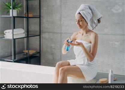Relaxed latina woman applying skincare cosmetics sitting on bath in modern bathroom. Female wrapped in towel squeezing cosmetic product from bottle for body care treatment after shower.. Woman apply natural skincare cosmetics sitting in modern bathroom. Body care treatment after shower
