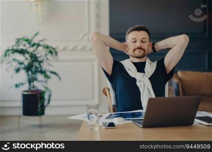Relaxed man freelancer or entrepreneur, wireless earphones, hands behind head, watching webinar on laptop, working from home.