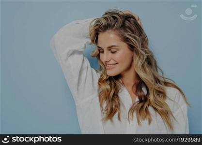 Relaxed romantic blonde woman with long wavy hair and hand behind her head, looking down with bright smile, dressed in white shirt with unbuttoned top while standing alone next to light blue wall. Relaxed blonde woman with hand behind her head