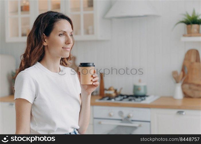 Relaxed smiling european woman with paper cup of coffee. Daily routine at home. Girl at kitchen in morning. Scandinavian stylish interior of apartment. White furniture, daylight.. Relaxed smiling woman with paper cup of coffee at home. Girl at kitchen in morning.