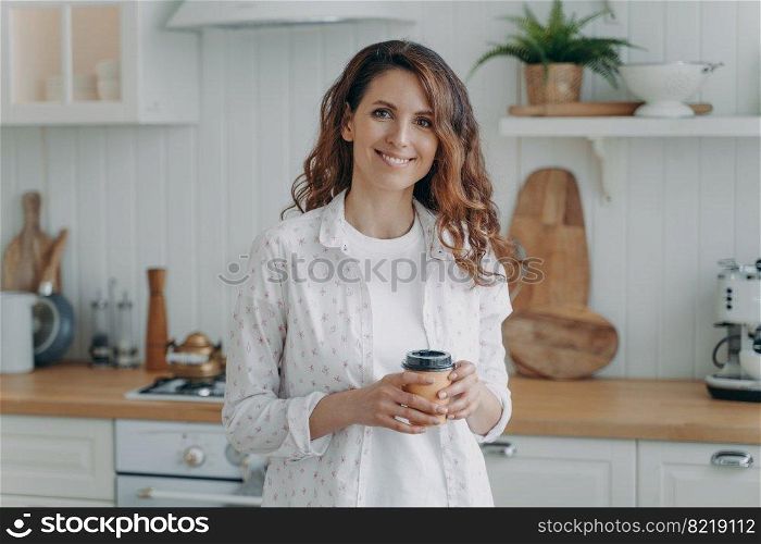 Relaxed smiling hispanic woman with paper cup of coffee standing in cozy modern kitchen, looking at camera. Pleased happy female homeowner or tenant housewife enjoying domestic routine at home.. Smiling hispanic woman with paper cup of coffee standing in cozy modern kitchen, looking at camera