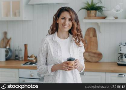 Relaxed smiling hispanic woman with paper cup of coffee standing in cozy modern kitchen, looking at camera. Pleased happy female homeowner or tenant housewife enjoying domestic routine at home.. Smiling hispanic woman with paper cup of coffee standing in cozy modern kitchen, looking at camera