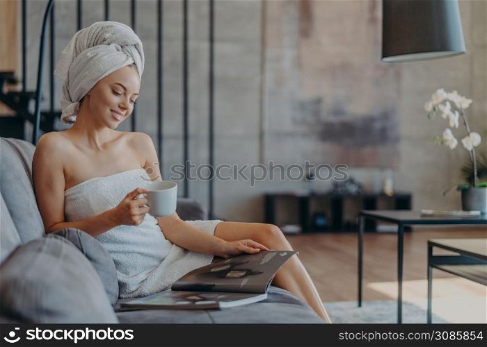Relaxed smiling young woman wrapped in towel after taking shower, drinks coffee and reads beauty magazine, sits onn comfortable sofa against cozy domestic interior. Wellness and beauty concept