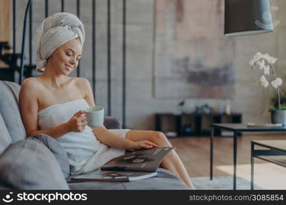 Relaxed smiling young woman wrapped in towel after taking shower, drinks coffee and reads beauty magazine, sits onn comfortable sofa against cozy domestic interior. Wellness and beauty concept