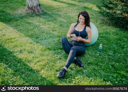 Relaxed sportswoman has rest after doing sport exercises with fitball, uses smartphone, listens favorite music in earphones from playlist, sits on green grass, bottle of fresh water near. Aerobics
