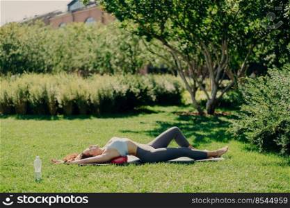 Relaxed sporty woman lying on fitness ball at karemat does aerobics exercises wears cropped top and leggings breathes fresh air deeply has perfect figure. People fitness and yoga practice concept