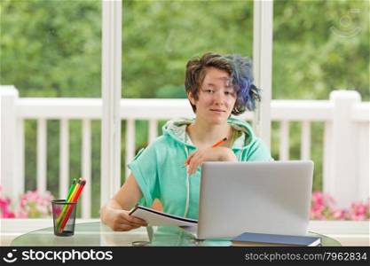 Relaxed teen girl, looking forward, holding pencil. Large windows in background with blurred out bright green trees and flowers.