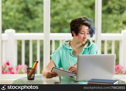 Relaxed teen girl taking notes while looking at computer. Large windows in background with blurred out bright green trees and flowers.