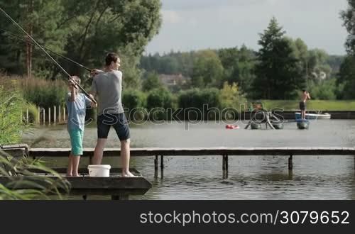 Relaxed teenage son and young dad casting spinning rod while angling together with fishing rod and reel on pond over rural landscape background. Side view. Fishermen with rod catching fish on the lake in summer. Slow motion.