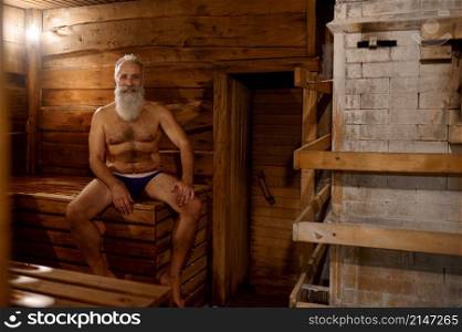 Relaxed thoughtful grey-haired senior man rest at bath house with oak broom. Wellness spa procedure. Grey-haired senior man rest at bath house