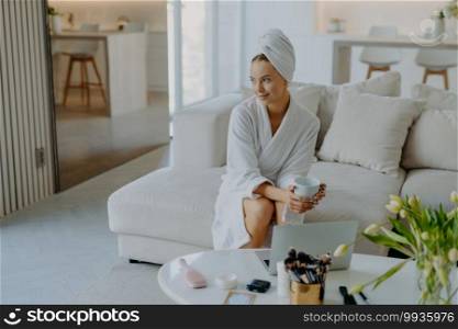 Relaxed thoughtful woman dressed in bathrobe and wrapped towel on head sits on sofa with cup of beverage near table with cosmetic products looks pensive aside poses against cozy home interior