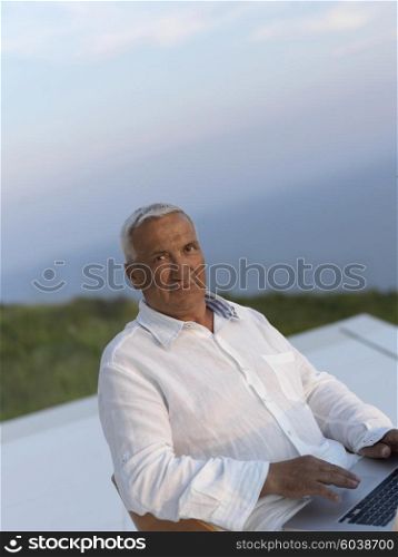 relaxed white hair senior man looking sunset from luxury balclony home and working on laptop computer
