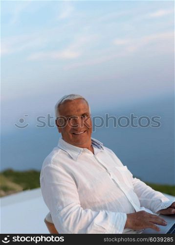 relaxed white hair senior man looking sunset from luxury balclony home and working on laptop computer