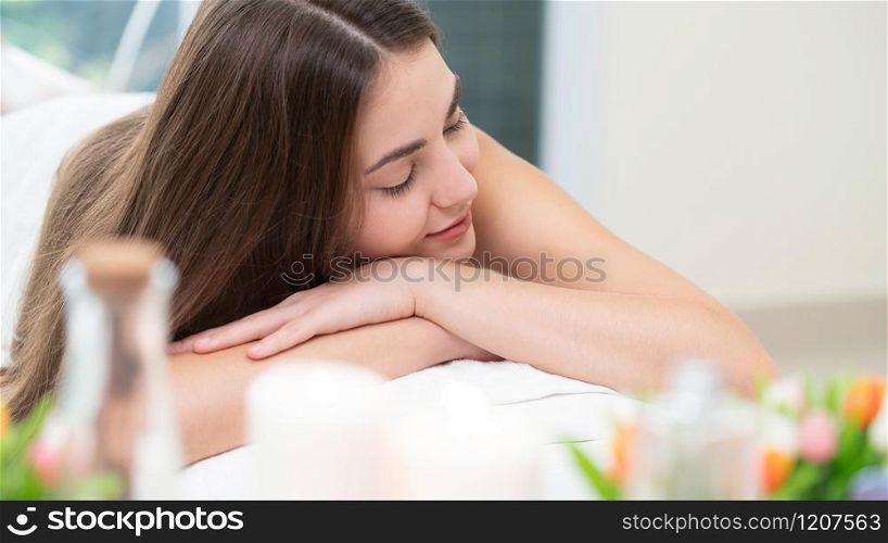 Relaxed woman lying on spa bed for aromatherapy massage in luxury spa with blurred foreground of spa treatment set including aromatic oil, candle and herbal scrub. Wellness and healing concept.. Woman lying on spa bed for massage in luxury spa.