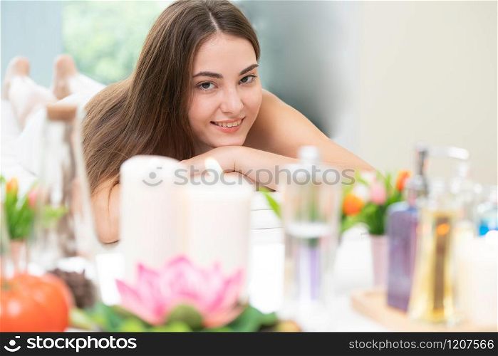 Relaxed woman lying on spa bed for aromatherapy massage in luxury spa with blurred foreground of spa treatment set including aromatic oil, candle and herbal scrub. Wellness and healing concept.. Woman lying on spa bed for massage in luxury spa.