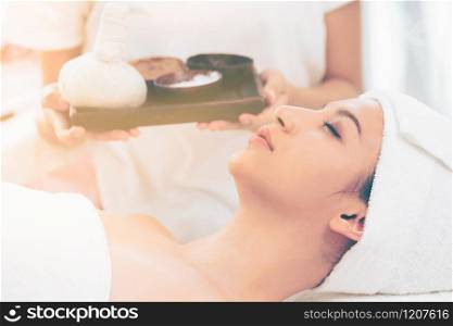 Relaxed woman lying on spa bed prepared for spa massage with therapist holding spa treatment set in background. Luxury wellness, stress relief and rejuvenation concept.