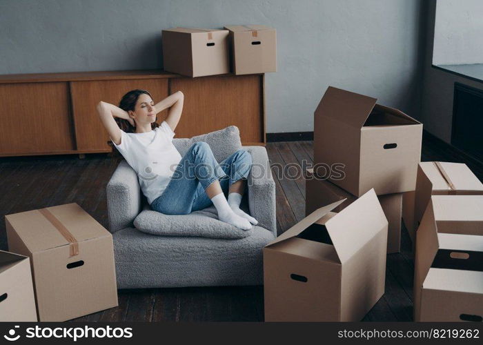 Relaxed woman sitting on armchair in empty house apartment with hands behind head, happy young female homeowner tenant relaxing during packing boxes on moving day to new home. Easy relocation concept.. Female sitting on armchair relaxes during packing boxes on moving day to new home. Easy relocation