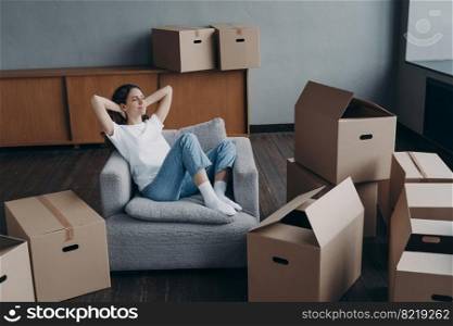 Relaxed woman sitting on armchair in empty house apartment with hands behind head, happy young female homeowner tenant relaxing during packing boxes on moving day to new home. Easy relocation concept.. Female sitting on armchair relaxes during packing boxes on moving day to new home. Easy relocation