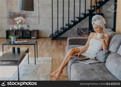 Relaxed woman with slender legs, healthy skin, enjoys drinking tea, sits on comfortable sofa, wrapped in bath towel, poses against cozy home interior. People, beauty, relaxation, lifestyle concept