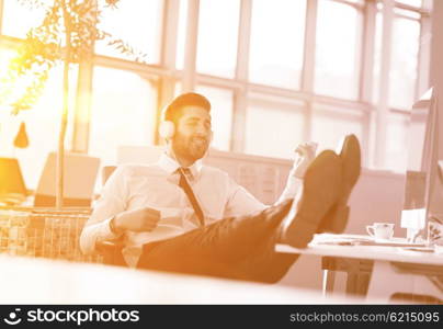 Relaxed young businessman first at workplace at early morning, listening music on headphones at modern startup office. Sunrise sun flare in background