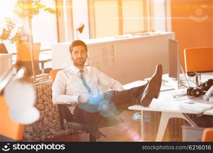 Relaxed young businessman first at workplace at early morning, listening music on headphones at modern startup office. Sunrise sun flare in background