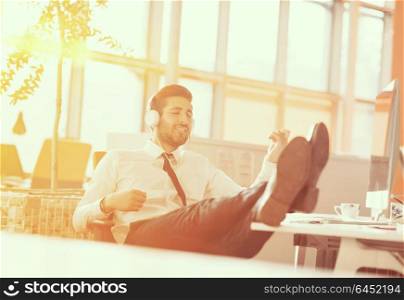 Relaxed young businessman first at workplace at early morning, listening music on headphones at modern startup office. Sunrise sun flare in background