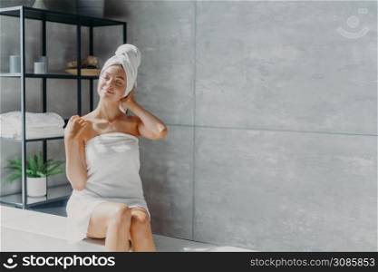 Relaxed young Caucasian female model wears towel wrapped on head, feels refreshed after taking shower, has healthy clean soft skin, poses in cozy bathroom. Women, beauty and hygiene concept.
