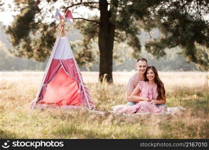 Relaxed young couple sitting near wigwam in park on sunny day. summer holiday vacation. man and woman are hugging outdoors and huving fun. Relaxed young couple sitting near wigwam in park on sunny day. summer holiday vacation. man and woman are hugging outdoors and huving fun.