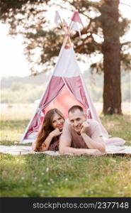 Relaxed young couple sitting near wigwam in park on sunny day. summer holiday vacation. man and woman are hugging outdoors and huving fun. Relaxed young couple sitting near wigwam in park on sunny day. summer holiday vacation. man and woman are hugging outdoors and huving fun.