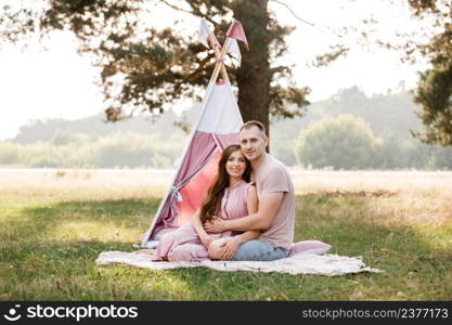 Relaxed young couple sitting near wigwam in park on sunny day. summer holiday vacation. man and woman are hugging outdoors and huving fun. Relaxed young couple sitting near wigwam in park on sunny day. summer holiday vacation. man and woman are hugging outdoors and huving fun.