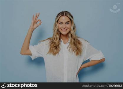 Relaxed young female showing ok gesture with hand and resting other one on her hip, assuring its alright and being confident it will all be fine while standing isolated next to blue wall. Relaxed blonde woman showing ok gesture with hand on hip