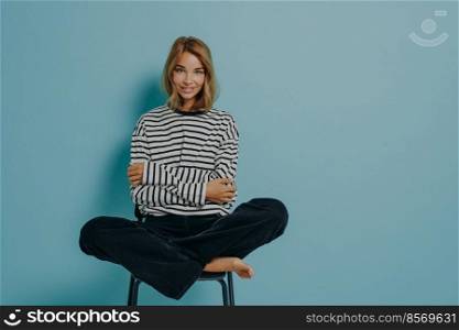 Relaxed young girl in striped shirt and jeans sitting with legs folded together in lotus pose on top of chair, huggs herself with hands pleasantly smiling, isolated on blue background with copy space. Relaxed young girl in striped shirt sitting with legs folded together in lotus pose on top of chair