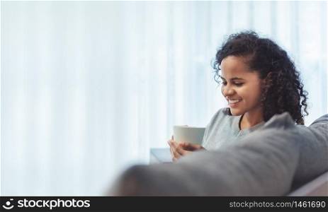 Relaxed Young smiling woman sitting on sofa while holding mug in winter morning. Attractive Hispanic female curly hair resting and enjoying in leisure time in living room at home. Carefree, copy space