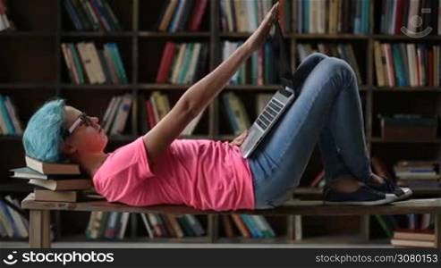 Relaxed young woman with blue hair and spectacles studying in library. Trendy hipster female student in casual comfortable outfit working on laptop while lying on the bench with head propped on a pile of books in university library.
