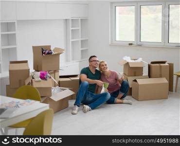 Relaxing in new house. Cheerful young couple sitting on the floor and drinking coffee while cardboard boxes laying all around them