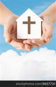 religion, christianity and charity concept - close up of woman hands holding paper house with christian cross symbol over blue sky with white cloud background