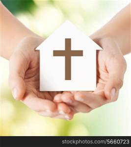 religion, christianity and charity concept - close up of woman hands holding paper house with christian cross symbol over green background