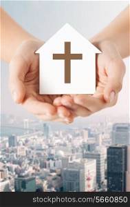 religion, christianity and charity concept - close up of woman hands holding paper house with christian cross symbol over city background