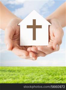 religion, christianity and charity concept - close up of woman hands holding paper house with christian cross symbol over blue sky with grass background