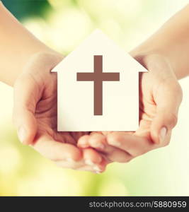 religion, christianity and charity concept - close up of woman hands holding paper house with christian cross symbol over green background