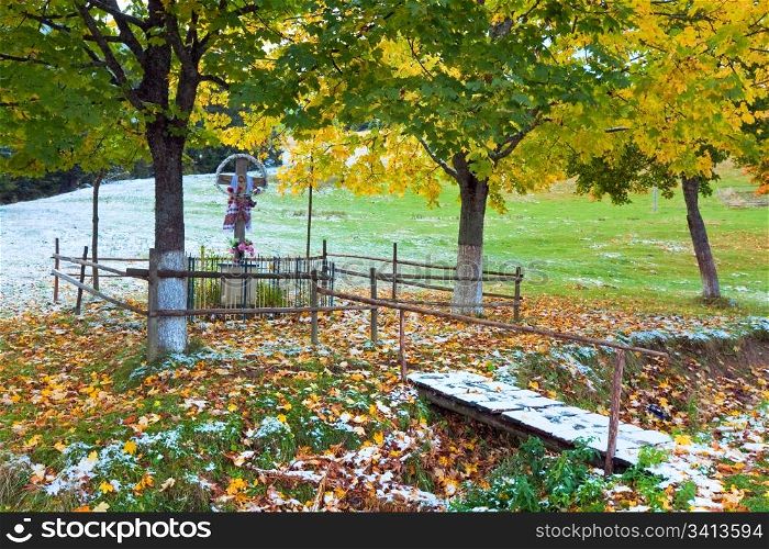 Religion christianity cross near road and first winter snow on autumn colourful maple foliage (Carpathian, Ukraine)