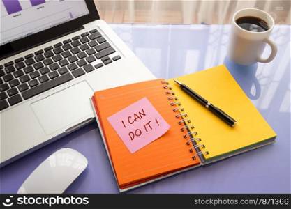 Reminder note on paper with message I CAN DO IT ! stick on colorful book with laptop and a cup of coffee on glass table, top view image