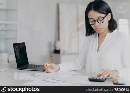 Remote accountant is calculating profit. Young hispanic bookkeeper in glasses and white blouse is working distantly from home. Female manager, serious girl is sitting at the desk and doing paperwork.. Remote accountant is calculating profit. Young hispanic bookkeeper is working distantly from home.