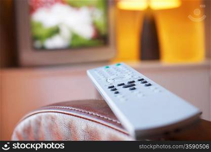 Remote control lying on armchair with television turned on in background close-up