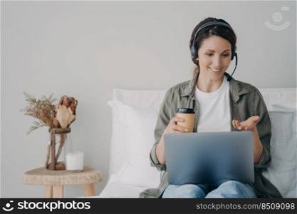 Remote education on quarantine. Young hispanic woman has group call on laptop at home. Freelancer has corporate internet session. Girl is talking in headphones having coffee in bedroom.. Remote education on quarantine. Young woman has group call on headphones having coffee.