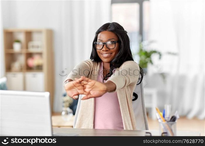 remote job, e-learning and people concept - happy smiling young african american woman in glasses with laptop computer stretching at home office. african american woman stretching at home office