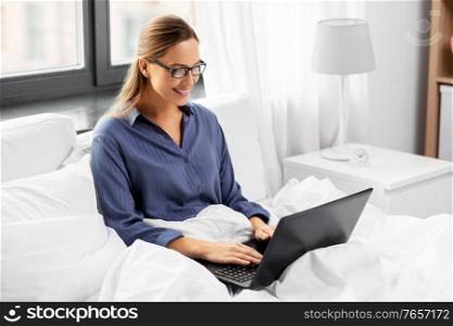remote job, people and business concept - young woman in glasses with laptop computer in bed working at home. young woman with laptop in bed at home bedroom