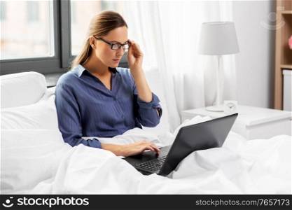 remote job, people and business concept - young woman in glasses with laptop computer in bed working at home. young woman with laptop in bed at home bedroom